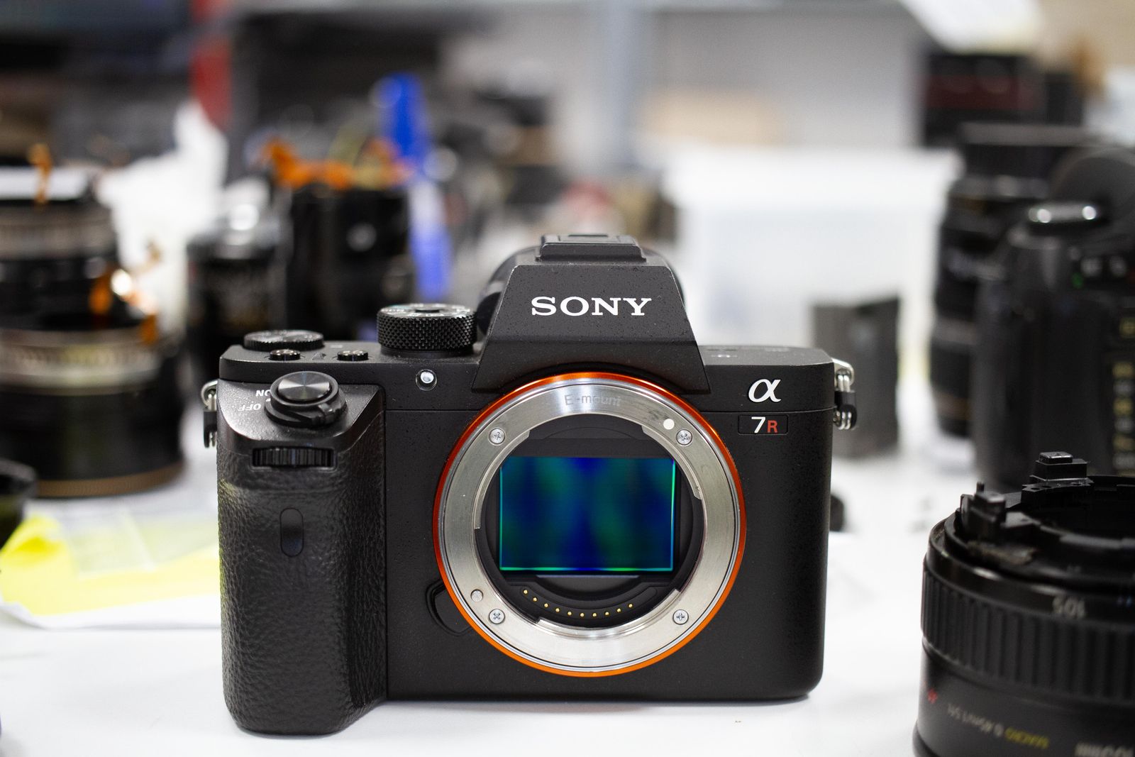 Sony A7R II con il sensore esposto sul banco del laboratorio Fotoriparatore, circondata da obiettivi in riparazione